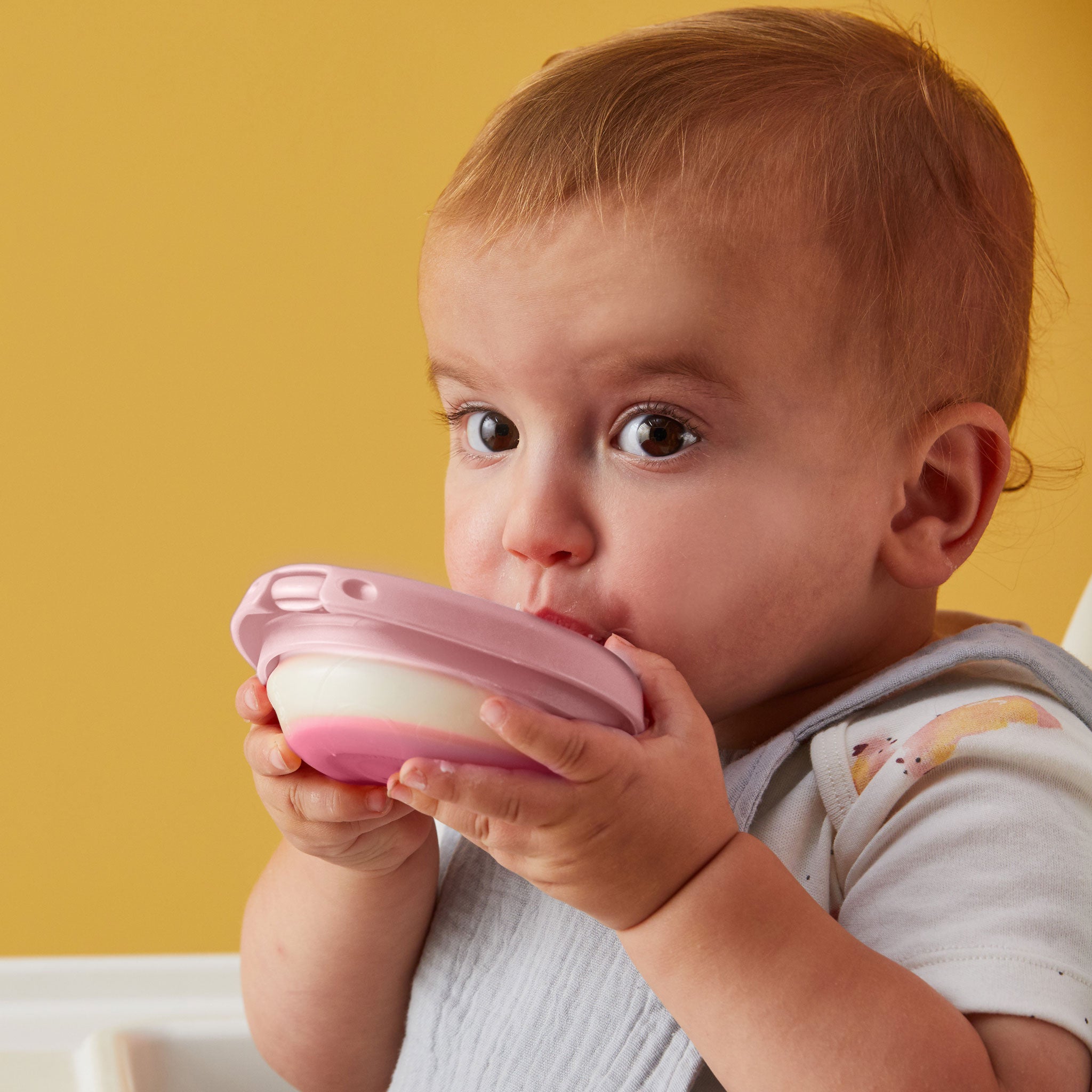 明るい黄色の背景の前にいる赤ちゃんが、ピンクの離乳食用容器を持って飲んでいる様子