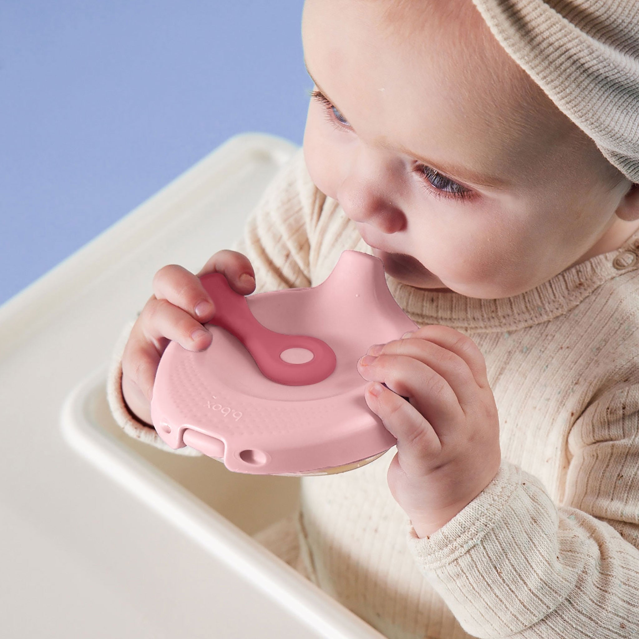 紫の背景の前にいる赤ちゃんが、ピンクの離乳食用容器を持って飲んでいる様子