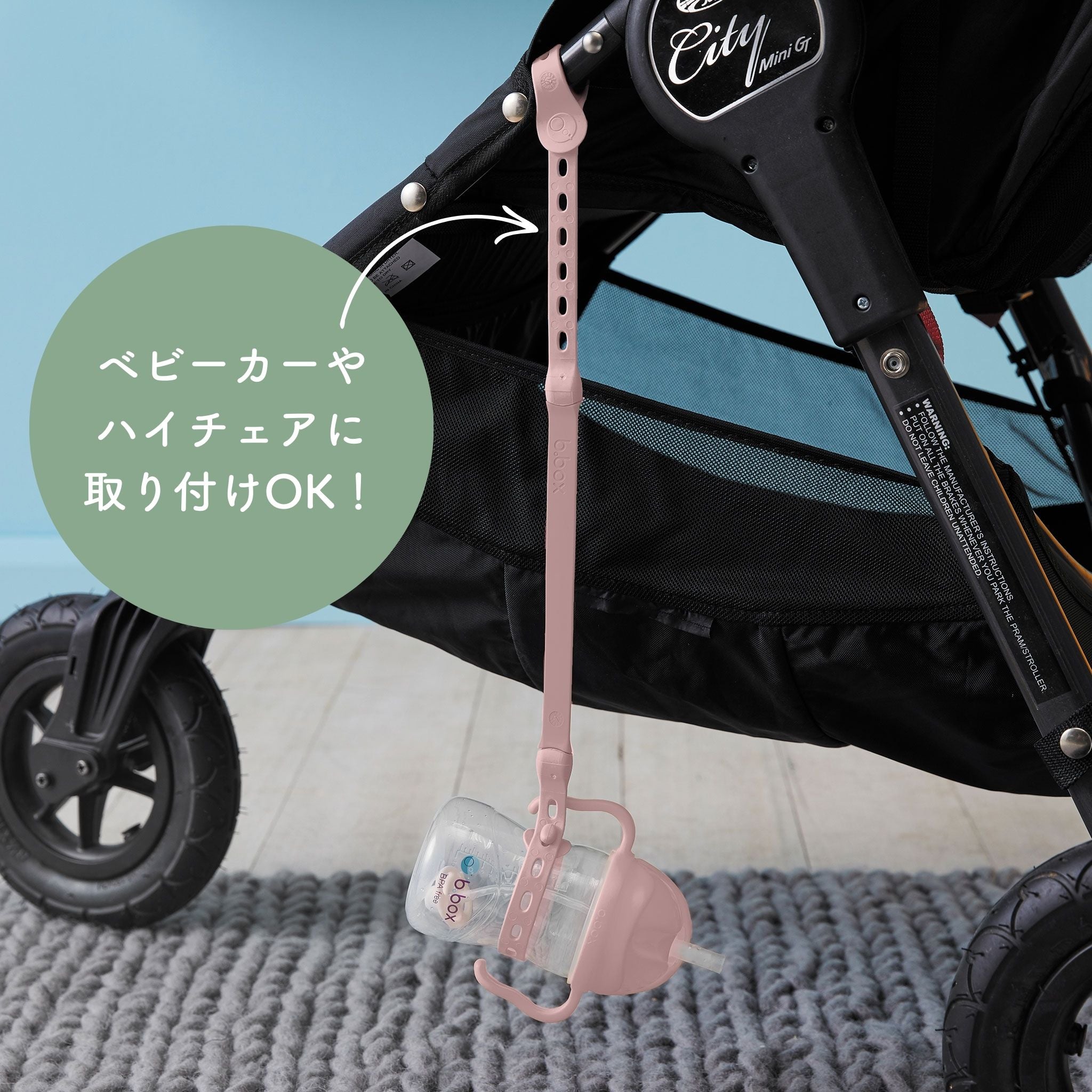 この画像は、ベビーカーのフレームに取り付けられたストラップに赤ちゃん用のドリンクボトルを固定している場面