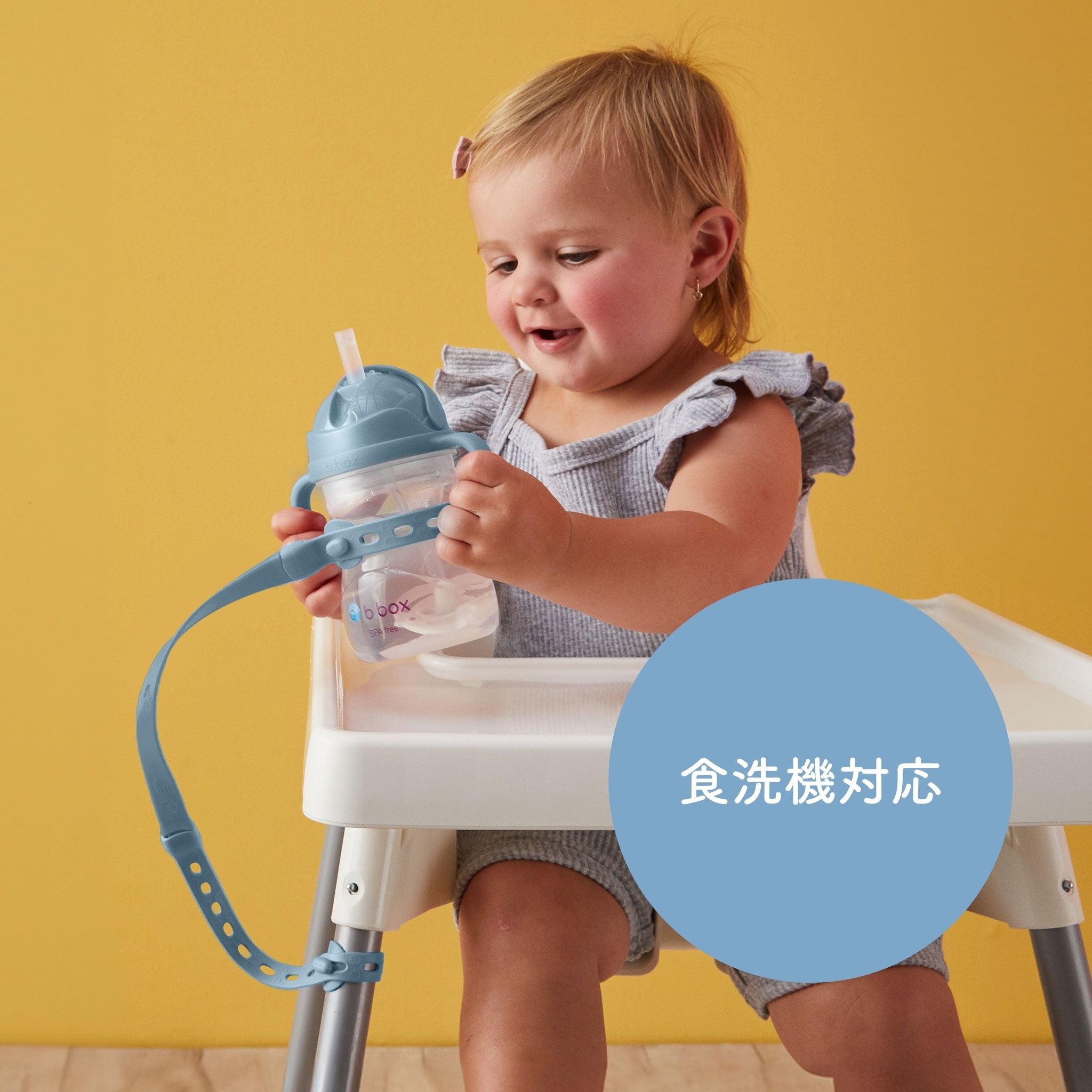 この画像には、小さな女の子が椅子に座ってストローボトルを持っている姿が写っています。ボトルにはシリコンストラップが付いており、椅子に固定されています。背景は黄色で、ボトルとストラップは青い色をしています。画像の右下に「食洗機対応」という文字がある青い円形のアイコンが表示されています。これは、このボトルが食器洗い機で洗えることを示しています。