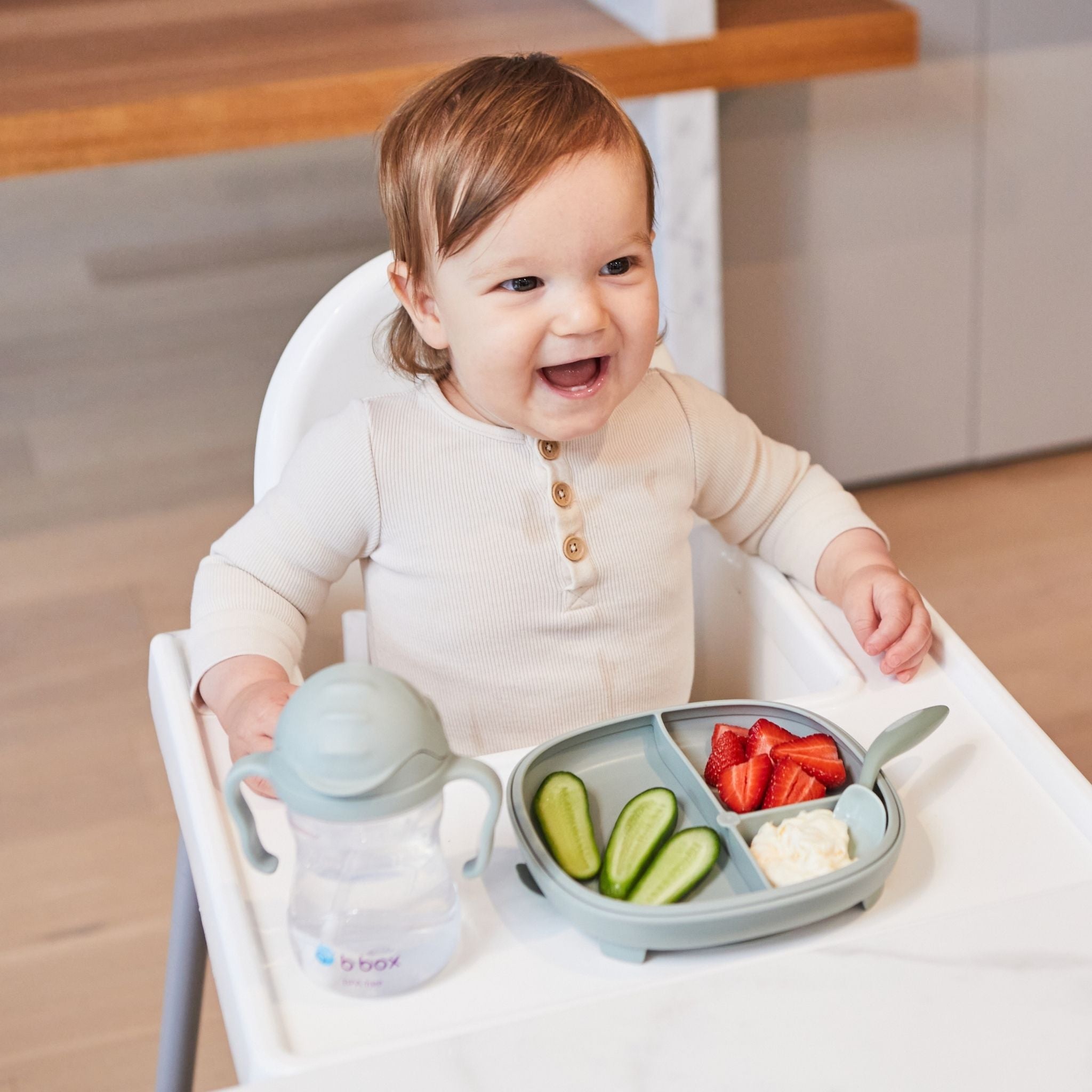 ハイチェアに座った赤ちゃんがフルーツと野菜が盛り付けられた幼児プレートとシッピーカップを使って食事を楽しんでいる様子