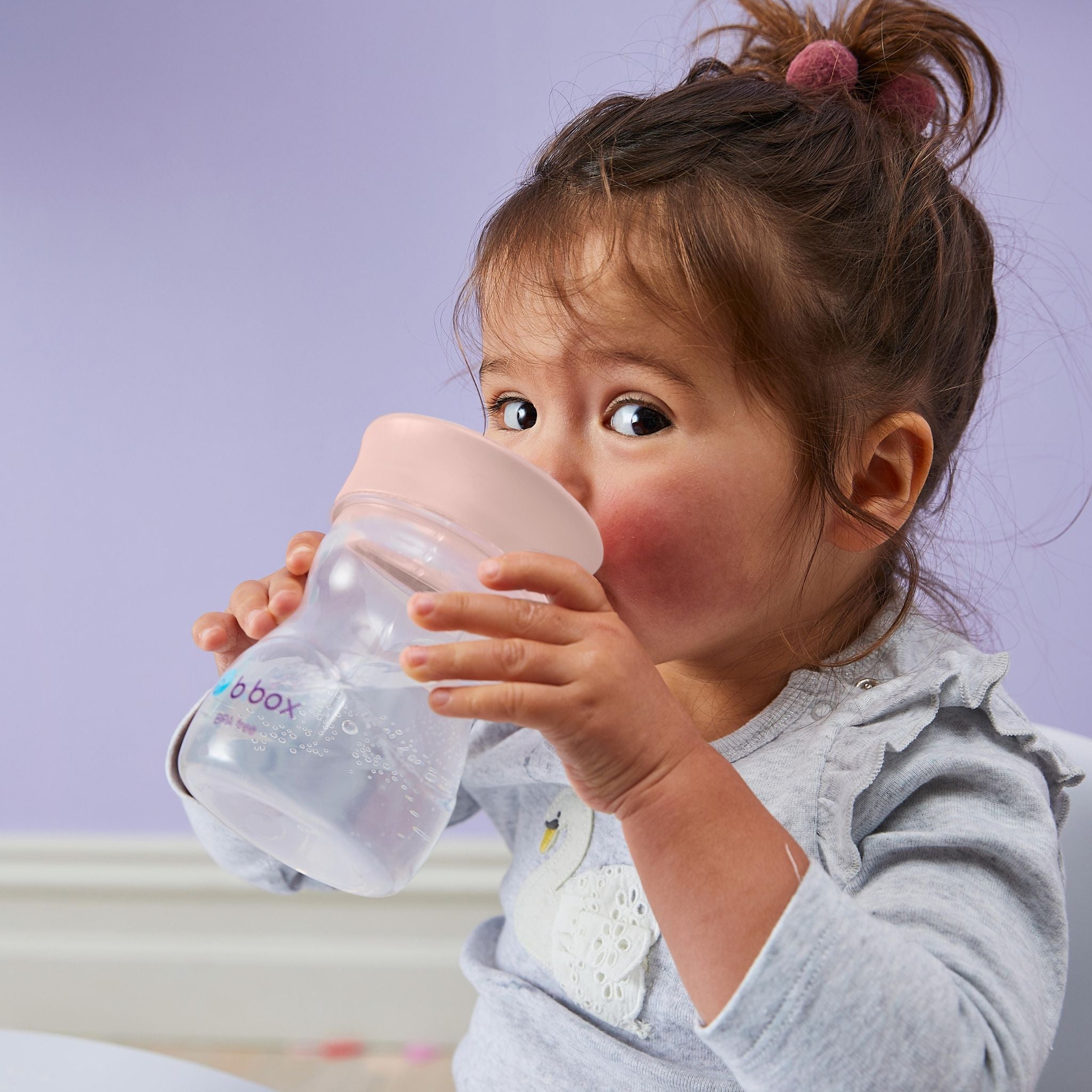 紫色の背景の前に女の子の赤ちゃんがコップ飲み練習アイテムで水分補給をしているところ